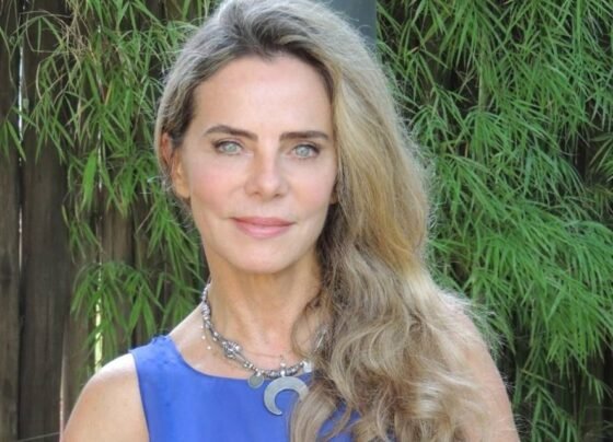 Foto de uma mulher loira e branca. Ela usa uma blusa azul, um colar grosso e prata. Ao fundo, mato verde - Metrópoles