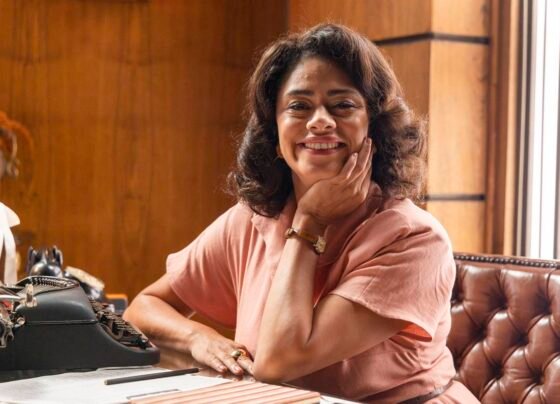 foto colorida de mulher negra com vestido rosa claro sentada e apoiada em cotovelo em cima da mesa - metrópoles